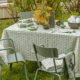 Une table de jardin présente la nappe verte et blanche XCOMM_NAPPE FORAL SAUGE 180X180, des serviettes de table assorties, des chaises vertes, des fleurs jaunes dans un vase, des bols, un chandelier vert et de la verdure en arrière-plan. la marque Toile de Jouy