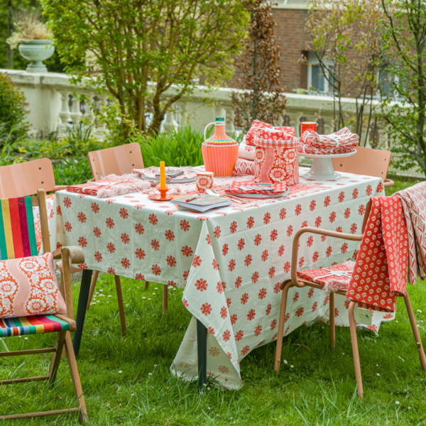 Une table de jardin est dressée pour un repas avec la nappe XCOMM_NAPPE FLORAL MANDARINE 180X180, entourée de quatre chaises colorées sur de l'herbe verte, avec des plantes et des arbres en arrière-plan. Des assiettes, des serviettes, une carafe et des tasses assorties sont posées sur la table. la marque Toile de Jouy
