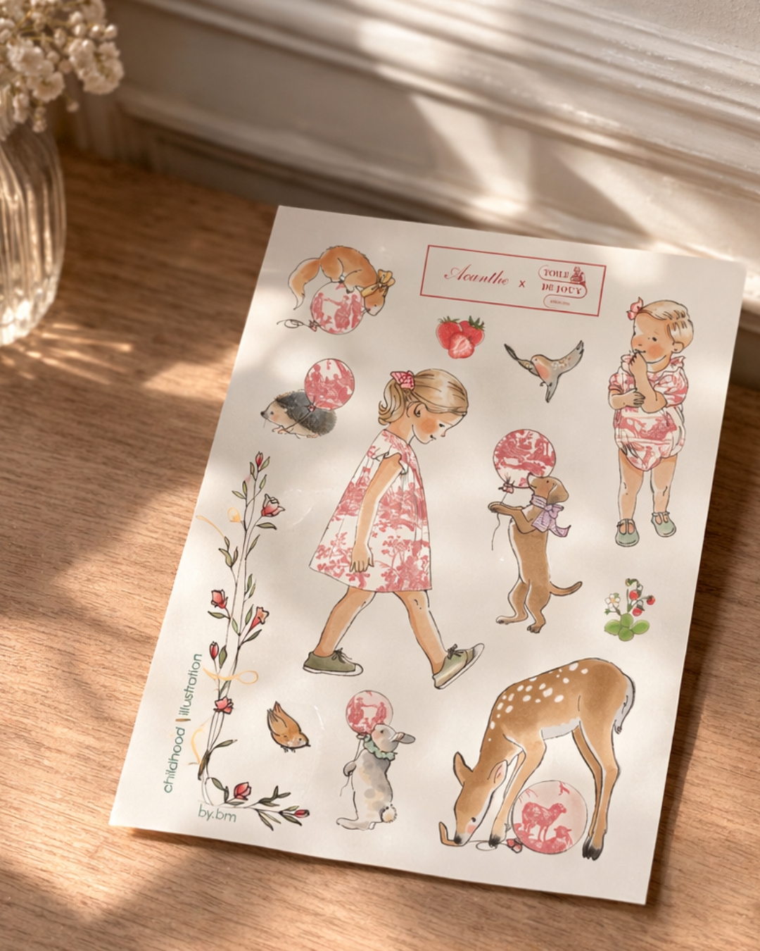 Une feuille d'autocollants sur une surface en bois présente PYJAMA SHORT FEMME "LES DELICES DES 4 SAISONS" ROSE- ACANTHE, représentant une jeune fille en robe rose à fleurs, un cerf, un chien et des motifs floraux avec la lumière du soleil qui projette des ombres douces. la marque Toile de Jouy