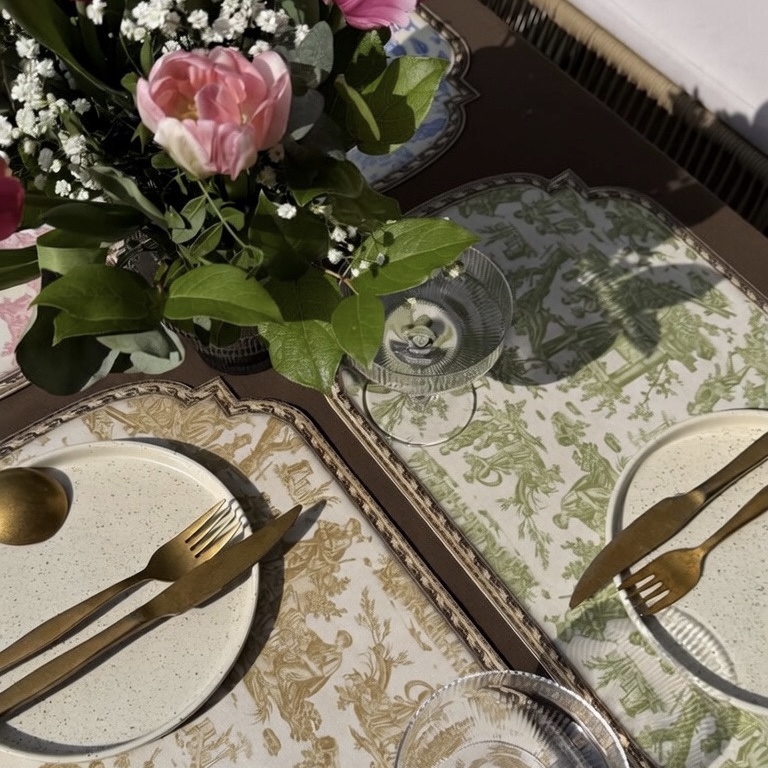 Gros plan sur une table dressée pour un repas, avec les sets de table SET DE TABLE VINYLE "OFFRANDE A L'AMOUR" SAFRAN, des ustensiles dorés, des assiettes mouchetées, des verres transparents et un centre de table en forme de bouquet de fleurs roses éclairé par la lumière du soleil. la marque Toile de Jouy