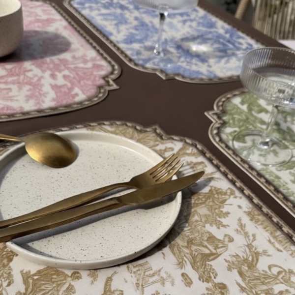 image7 Gros plan d'un service de table composé d'une assiette blanche mouchetée, d'une cuillère, d'un couteau et d'une fourchette dorés sur un set de table à motifs beiges, avec d'autres sets de table de couleur pastel et des verres vides à l'arrière-plan. la marque Toile de Jouy