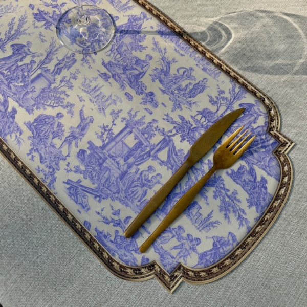 Un verre à vin, une fourchette et un couteau dorés sont posés sur le SET DE TABLE VINYLE "OFFRANDE A L'AMOUR" SAFRAN, qui présente des scènes pastorales bleues et une bordure ornée sur une nappe légèrement texturée. la marque Toile de Jouy