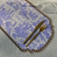 Un verre à vin, une fourchette et un couteau dorés sont posés sur le SET DE TABLE VINYLE "OFFRANDE A L'AMOUR" SAFRAN, qui présente des scènes pastorales bleues et une bordure ornée sur une nappe légèrement texturée. la marque Toile de Jouy