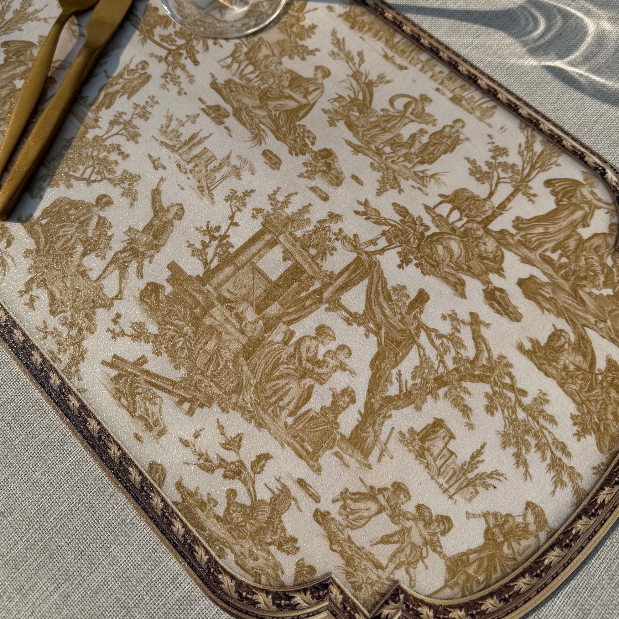Gros plan sur le SET DE TABLE VINYLE "OFFRANDE A L'AMOUR" SAFRAN, représentant des scènes pastorales brunes détaillées, présenté sur une nappe claire avec des couverts dorés et un verre partiellement visible sur la gauche. la marque Toile de Jouy