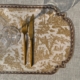 Une fourchette et un couteau dorés reposent sur le SET DE TABLE VINYLE "OFFRANDE A L'AMOUR" SAFRAN, à côté d'un verre à vin vide sur une nappe beige texturée. la marque Toile de Jouy