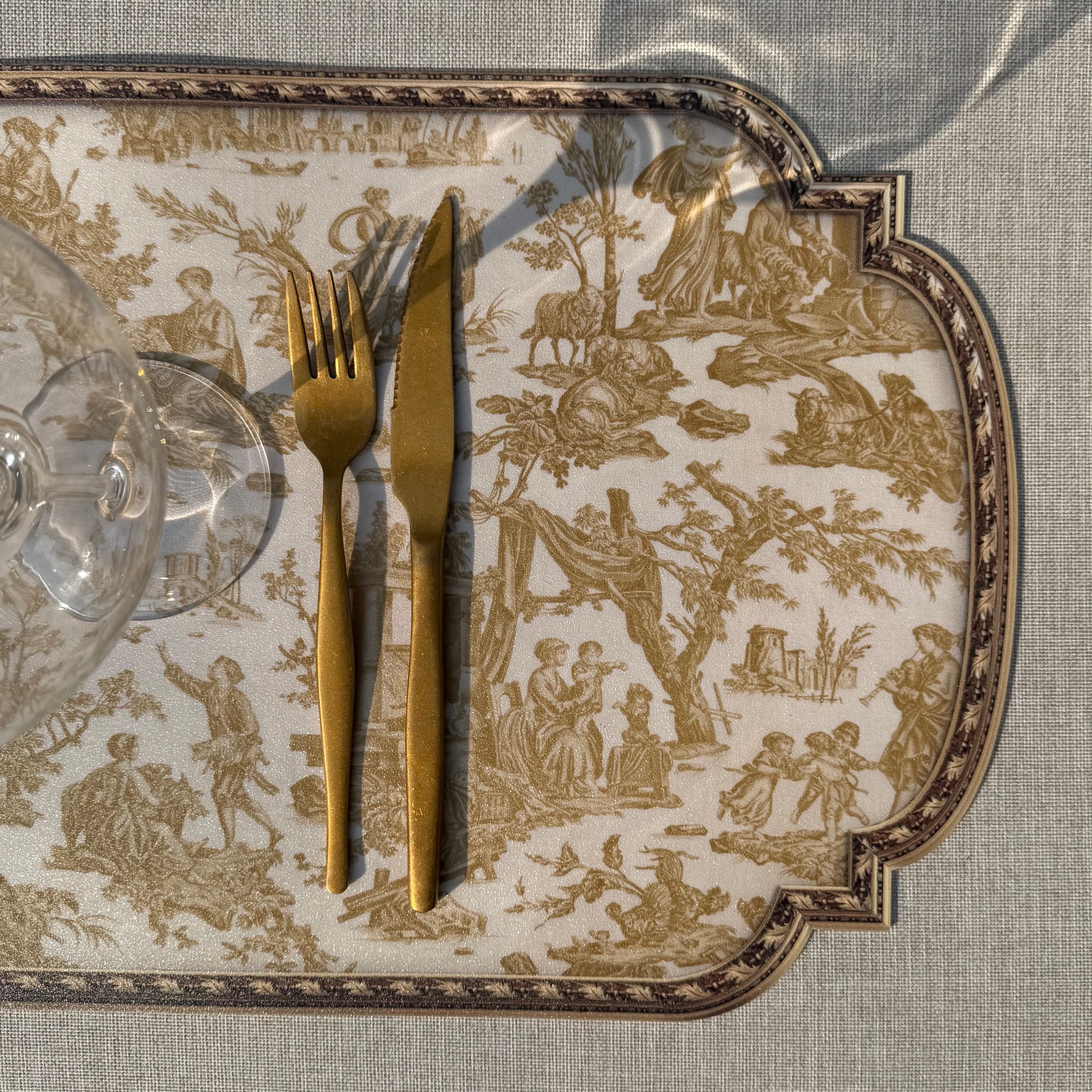 Une fourchette et un couteau dorés reposent sur le SET DE TABLE VINYLE "OFFRANDE A L'AMOUR" SAFRAN, à côté d'un verre à vin vide sur une nappe beige texturée. la marque Toile de Jouy