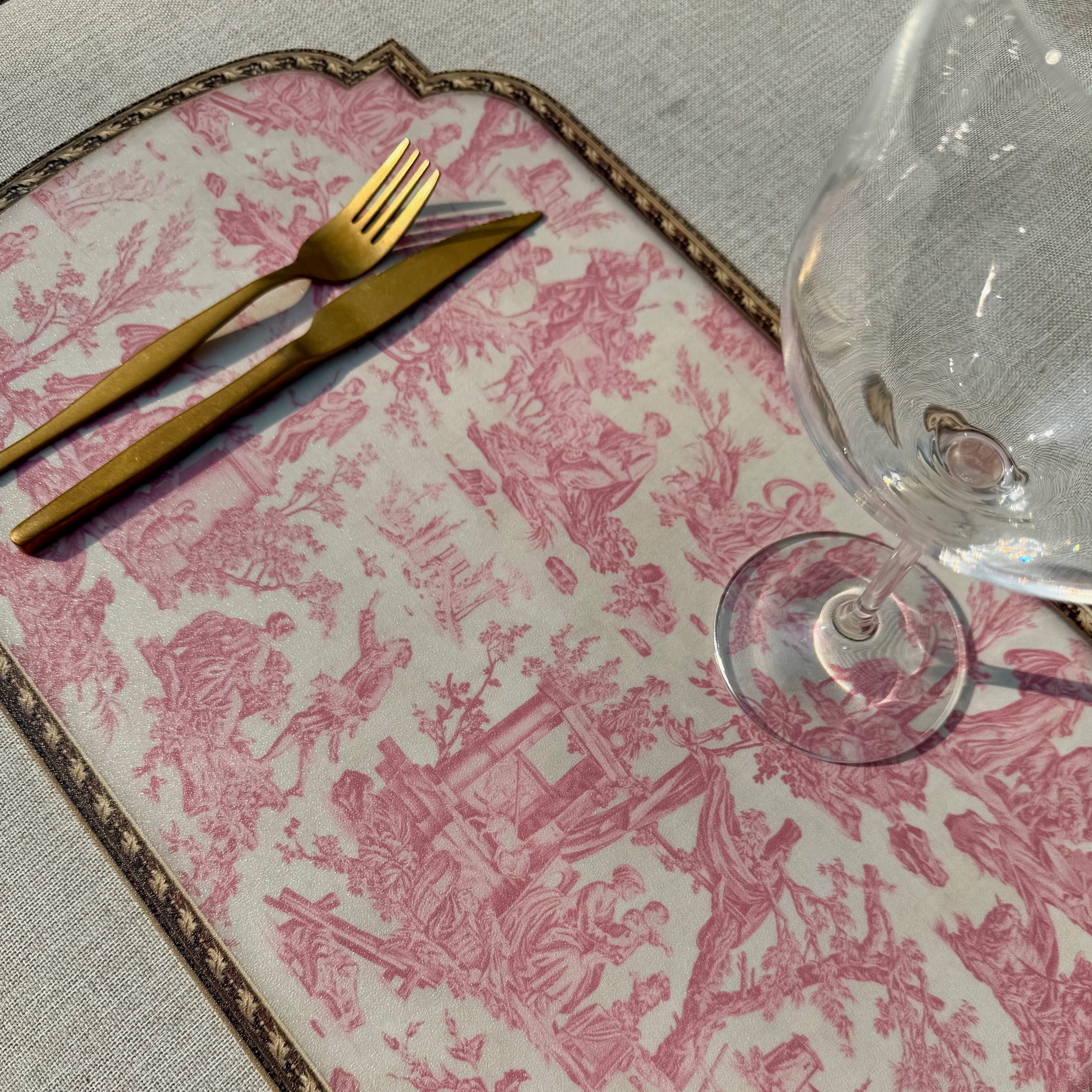 Une fourchette et un couteau dorés reposent sur le SET DE TABLE VINYLE "OFFRANDE A L'AMOUR" SAFRAN, orné de motifs roses et blancs. Un verre à vin vide est posé à droite sur une nappe claire. la marque Toile de Jouy