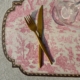 Une fourchette et un couteau dorés reposent en diagonale sur le set de table en vinyle Safran "OFFRANDE A L'AMOUR" aux motifs classiques roses et blancs, à côté de la base d'un verre transparent. la marque Toile de Jouy