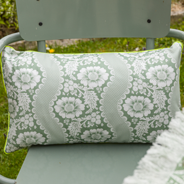Le XCOMM_COUSSIN 30X50 FLEURS SAUGE, un coussin rectangulaire avec un motif floral vert et blanc, est posé sur une chaise en métal vert clair à l'extérieur, entourée d'herbe. la marque Toile de Jouy