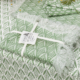 Un ensemble de deux sets de table à franges vertes et blanches, "XCOMM_SET DE TABLE/2 EPIS SAUGE", présente un motif feuillu, noué par un ruban crème et présenté sur un fond assorti avec un autre textile visible. la marque Toile de Jouy