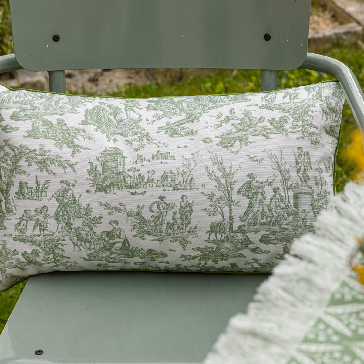 Le XCOMM_COUSSIN 30X50 FLEURS SAUGE, un coussin rectangulaire aux motifs floraux de sauge, repose en plein air sur une chaise en métal vert clair, partiellement ombragée par un tissu à franges. la marque Toile de Jouy