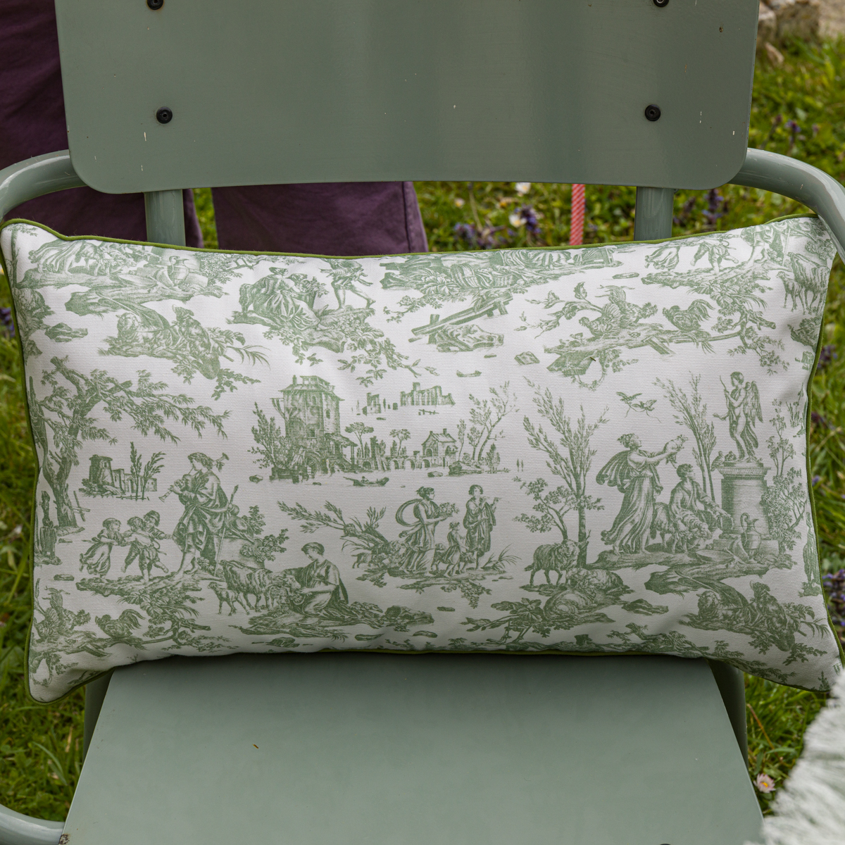 Le XCOMM_COUSSIN 30X50 FLEURS SAUGE est un coussin rectangulaire représentant des scènes de campagne en toile verte sur tissu blanc, en extérieur sur une chaise en métal vert clair sur fond d'herbe. la marque Toile de Jouy