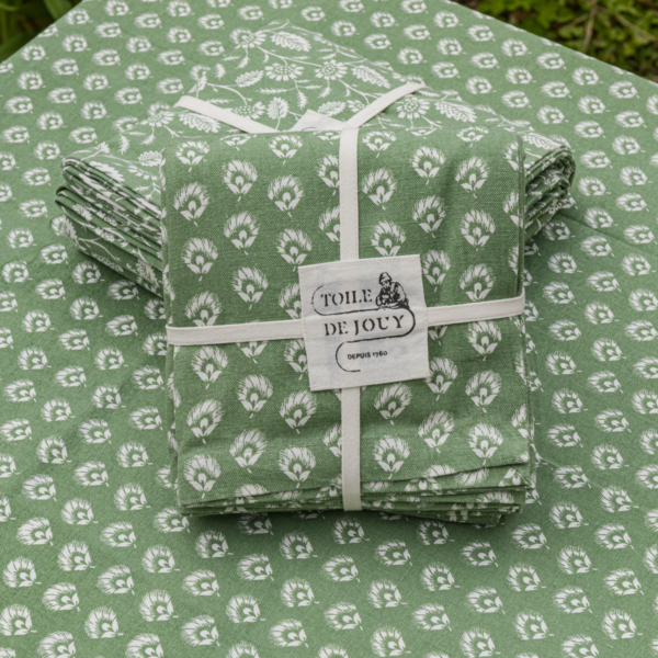 Le XCOMM_NAPPE CHARDON SAGE 150X150 présente un tissu vert plié avec un motif floral blanc attaché avec un ruban blanc étiqueté "Toile de Jouy Depuis 1760", avec un tissu assorti empilé en dessous et à l'arrière-plan. la marque Toile de Jouy