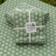 Le XCOMM_NAPPE CHARDON SAGE 150X150 présente un tissu vert plié avec un motif floral blanc attaché avec un ruban blanc étiqueté "Toile de Jouy Depuis 1760", avec un tissu assorti empilé en dessous et à l'arrière-plan. la marque Toile de Jouy
