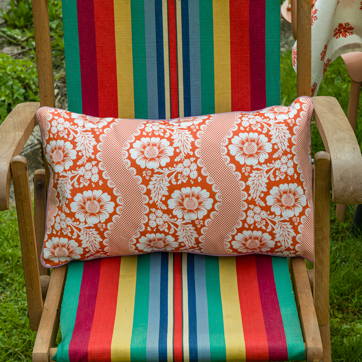 Une chaise en bois avec une assise et un dossier en tissu rayé coloré tient le XCOMM_COUSSIN 30X50 FLEURS MANDARINE, un coussin rectangulaire orange avec des motifs floraux blancs. La chaise est posée sur de l'herbe verte en plein air. la marque Toile de Jouy