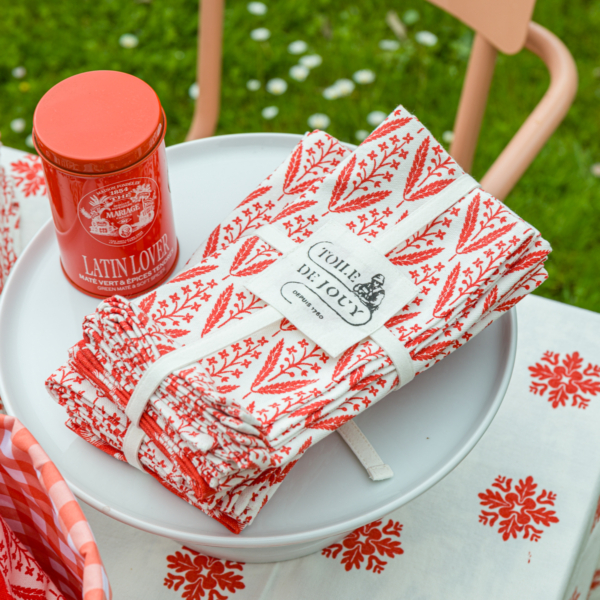 Un paquet de serviettes XCOMM_SERVIETTES/6 CHARDON MANDARINE est posé sur une assiette blanche à côté d'une boîte rouge étiquetée "Latin Lover", avec en arrière-plan un panier à carreaux rouges et blancs, une nappe fleurie et de l'herbe avec des marguerites. la marque Toile de Jouy