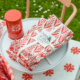 Un paquet de serviettes XCOMM_SERVIETTES/6 CHARDON MANDARINE est posé sur une assiette blanche à côté d'une boîte rouge étiquetée "Latin Lover", avec en arrière-plan un panier à carreaux rouges et blancs, une nappe fleurie et de l'herbe avec des marguerites. la marque Toile de Jouy