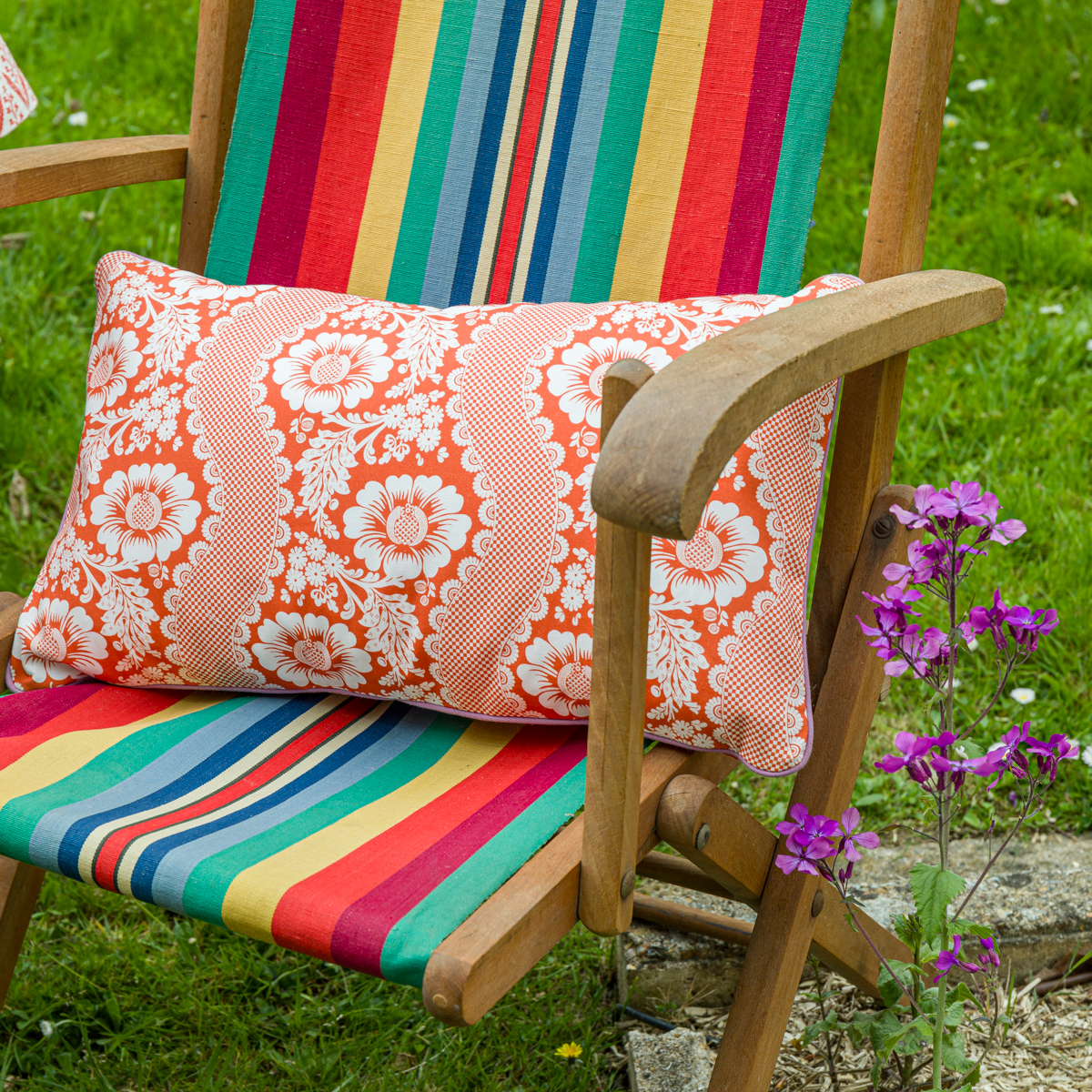 Une chaise en bois avec un siège à rayures colorées et l'oreiller XCOMM_COUSSIN 30X50 FLEURS MANDARINE est posée sur de l'herbe à côté de fleurs violettes. la marque Toile de Jouy