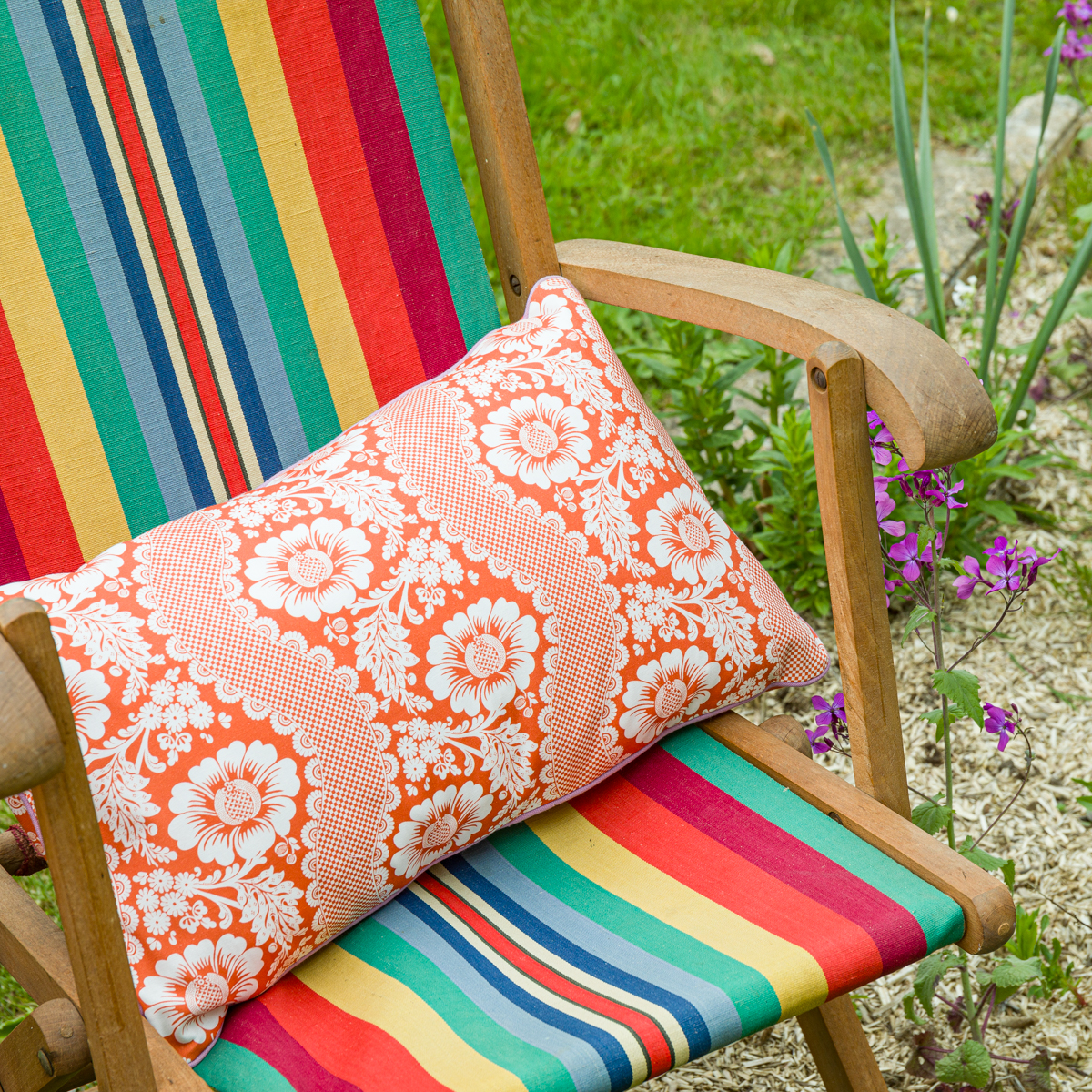 Une chaise de jardin en bois avec un tissu à rayures vives porte le XCOMM_COUSSIN 30X50 FLEURS MANDARINE, un coussin orange et blanc à motifs floraux. Des fleurs violettes s'épanouissent à proximité, dans une petite plate-bande. la marque Toile de Jouy