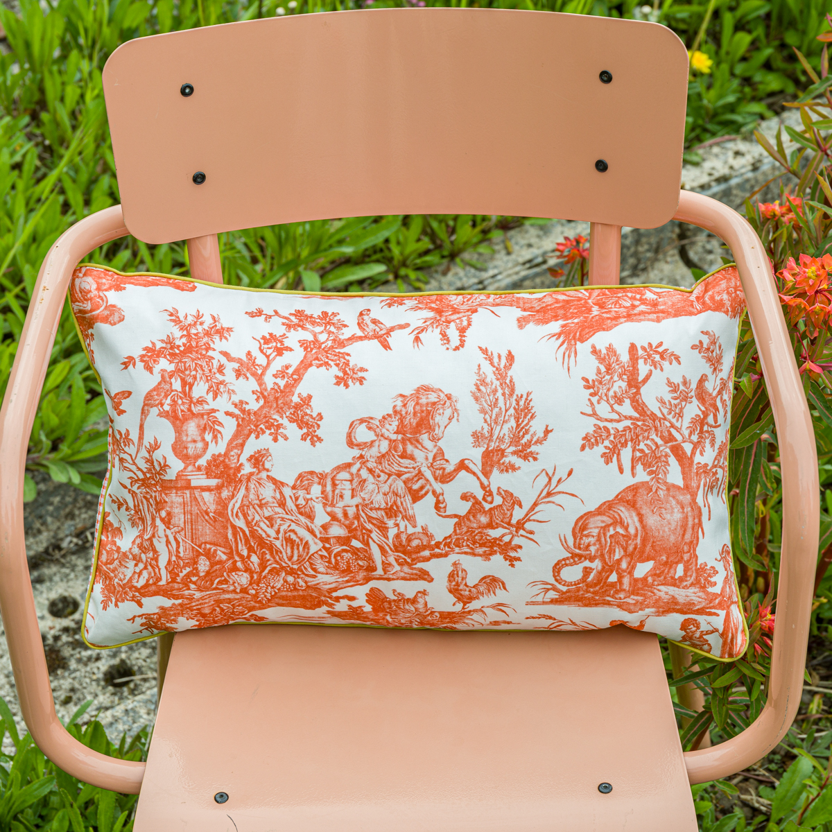 Une chaise en métal rose avec le XCOMM_COUSSIN 30X50 FLEURS MANDARINE, un coussin rectangulaire avec un imprimé floral orange et blanc, placé à l'extérieur avec de l'herbe verte et des fleurs en arrière-plan. la marque Toile de Jouy