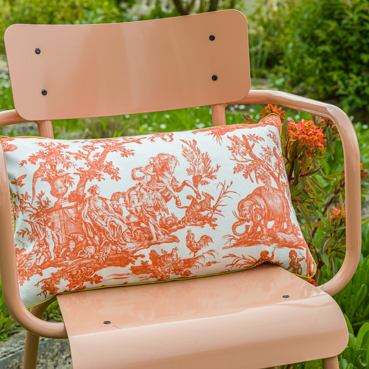 L'oreiller XCOMM_COUSSIN 30X50 FLEURS MANDARINE, qui présente un motif de toile orange et blanche, est posé sur une chaise en métal de couleur pêche, en plein air, au milieu d'une verdure luxuriante et de fleurs orange éclatantes. la marque Toile de Jouy