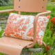 Une chaise en métal de couleur pêche présente à l'extérieur l'oreiller XCOMM_COUSSIN 30X50 FLEURS MANDARINE avec un motif pastoral orange et blanc sur l'assise, avec de l'herbe verte et des fleurs orange en arrière-plan. la marque Toile de Jouy