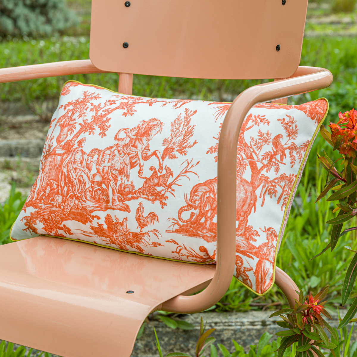 Une chaise en métal de couleur pêche présente à l'extérieur l'oreiller XCOMM_COUSSIN 30X50 FLEURS MANDARINE avec un motif pastoral orange et blanc sur l'assise, avec de l'herbe verte et des fleurs orange en arrière-plan. la marque Toile de Jouy
