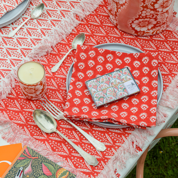 Un décor de table festif présente le XCOMM_SET DE TABLE /2 EPIS MANDARINE, avec de l'argenterie vintage, une bougie, une serviette à motifs rouges sur une assiette et une boîte de conserve décorative, le tout sur une nappe à franges rouge et blanche. la marque Toile de Jouy