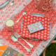 Un décor de table festif présente le XCOMM_SET DE TABLE /2 EPIS MANDARINE, avec de l'argenterie vintage, une bougie, une serviette à motifs rouges sur une assiette et une boîte de conserve décorative, le tout sur une nappe à franges rouge et blanche. la marque Toile de Jouy