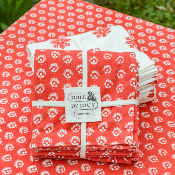 XCOMM_NAPPE CHARDON MANDARINE 150X150 est posée sur la table, avec des serviettes à fleurs rouges et blanches nouées par un ruban blanc et portant l'étiquette "Toile de Jouy", ainsi que des serviettes blanches pliées présentant un motif similaire. la marque Toile de Jouy