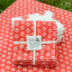 XCOMM_NAPPE CHARDON MANDARINE 150X150 est posée sur la table, avec des serviettes à fleurs rouges et blanches nouées par un ruban blanc et portant l'étiquette "Toile de Jouy", ainsi que des serviettes blanches pliées présentant un motif similaire. la marque Toile de Jouy