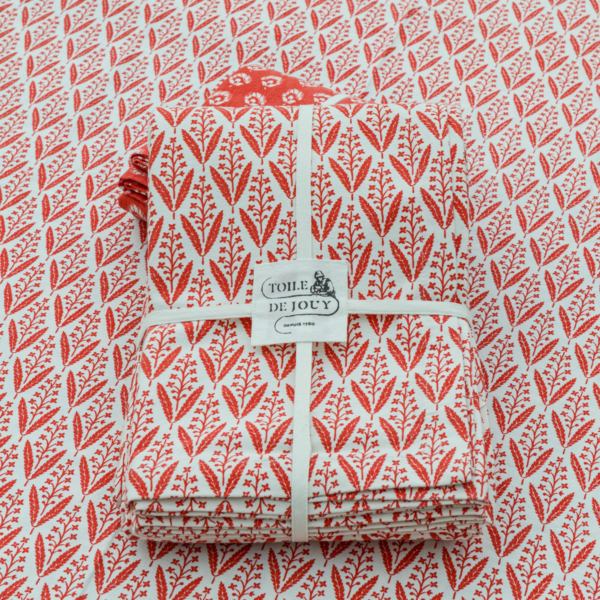 Une pile de serviettes blanches pliées avec un motif de feuilles rouges, nouées avec un ruban "Toile de Jouy", repose sur un fond de tissu XCOMM_NAPPE FLORAL MANDARINE 180X180 assorti. la marque Toile de Jouy