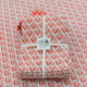 Une pile de serviettes blanches pliées avec un motif de feuilles rouges, nouées avec un ruban "Toile de Jouy", repose sur un fond de tissu XCOMM_NAPPE FLORAL MANDARINE 180X180 assorti. la marque Toile de Jouy