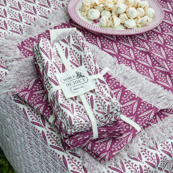 Un ensemble de serviettes XCOMM_SERVIETTES/6 CHARDON AMARANTE à franges violettes et blanches, nouées avec un ruban, est posé sur une nappe assortie. À côté, une assiette de petits biscuits meringués blancs. la marque Toile de Jouy