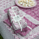 Un ensemble de serviettes XCOMM_SERVIETTES/6 CHARDON AMARANTE à franges violettes et blanches, nouées avec un ruban, est posé sur une nappe assortie. À côté, une assiette de petits biscuits meringués blancs. la marque Toile de Jouy