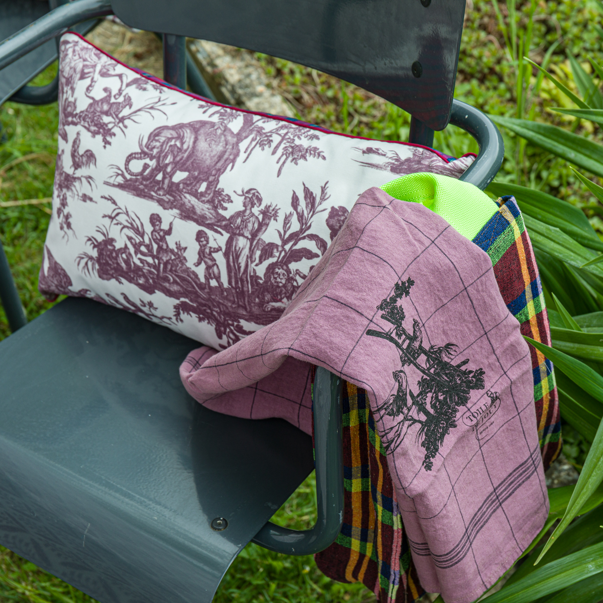 À l'extérieur, une chaise métallique grise supporte le coussin décoratif XCOMM_COUSSIN 30X50 FLEURS SAUGE, ainsi qu'un tissu écossais rose plié, entouré d'herbe et de plantes vertes. la marque Toile de Jouy