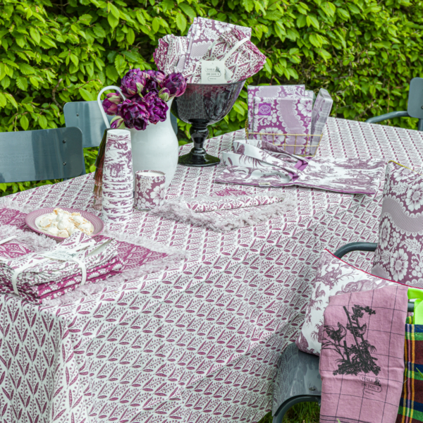 Une table de jardin est ornée de la nappe violette et blanche XCOMM_NAPPE EPIS AMARANTE 180X270, de serviettes, d'assiettes, de tasses et de décorations assorties. Un vase contenant des tulipes violettes est posé sur la table, encadré par des buissons verts et feuillus en arrière-plan. la marque Toile de Jouy