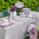 Une table de jardin est ornée de la nappe violette et blanche XCOMM_NAPPE EPIS AMARANTE 180X270, de serviettes, d'assiettes, de tasses et de décorations assorties. Un vase contenant des tulipes violettes est posé sur la table, encadré par des buissons verts et feuillus en arrière-plan. la marque Toile de Jouy