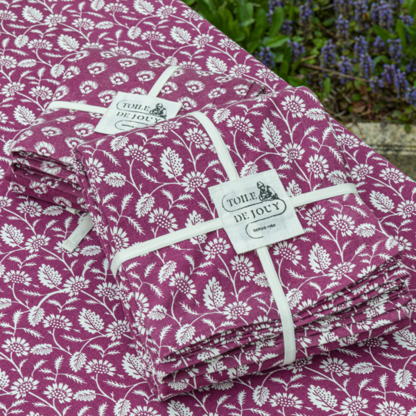 Des liasses pliées de XCOMM_NAPPE FLORAL AMARANTE 180X180, en tissu violet avec des motifs floraux blancs et nouées avec un ruban blanc, sont présentées sur un tissu assorti avec de la verdure et des fleurs bleues dans le coin. la marque Toile de Jouy