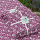 Des liasses pliées de XCOMM_NAPPE FLORAL AMARANTE 180X180, en tissu violet avec des motifs floraux blancs et nouées avec un ruban blanc, sont présentées sur un tissu assorti avec de la verdure et des fleurs bleues dans le coin. la marque Toile de Jouy