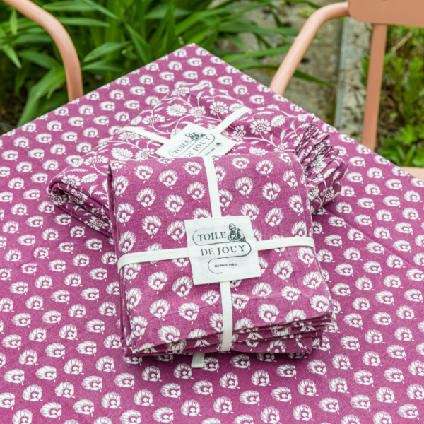 Deux paquets de tissu pliés avec un motif floral blanc sur fond violet, noués avec un ruban blanc et étiquetés "Toile de Jouy", sont placés sur la nappe XCOMM_NAPPE CHARDON AMARANTE 150X150 à l'extérieur, près d'un jardin. la marque Toile de Jouy