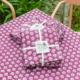 Deux paquets de tissu pliés avec un motif floral blanc sur fond violet, noués avec un ruban blanc et étiquetés "Toile de Jouy", sont placés sur la nappe XCOMM_NAPPE CHARDON AMARANTE 150X150 à l'extérieur, près d'un jardin. la marque Toile de Jouy