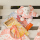 La trousse XCOMM_TROUSSE PENELOPE OFFRANDE PEACH PINK, accompagnée de crayons de couleur présentés dans une boîte en bois, d'un livre de coloriage et d'articles en tissu ornés de motifs roses, est disposée sur une surface en bois blanc. la marque Toile de Jouy