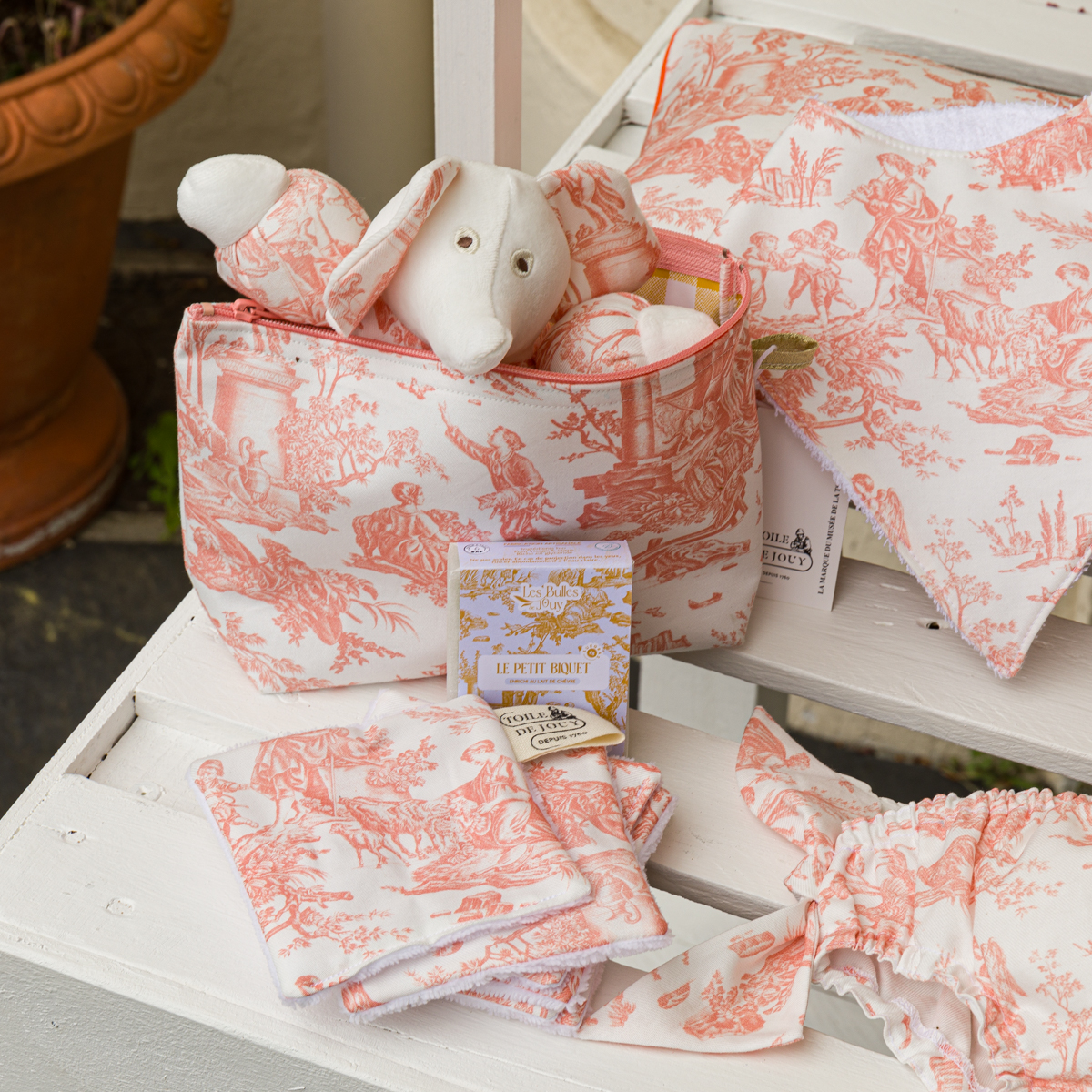 Le coffret cadeau « XCOMM_TROUSSE PENELOPE OFFRANDE PEACH PINK », orné d’un motif de toile de Jouy rose et blanc, comprend une pochette à fermeture éclair contenant un lapin en peluche, un oreiller, des lingettes et une boîte de savon, le tout joliment disposé sur un plateau en bois blanc. la marque Toile de Jouy