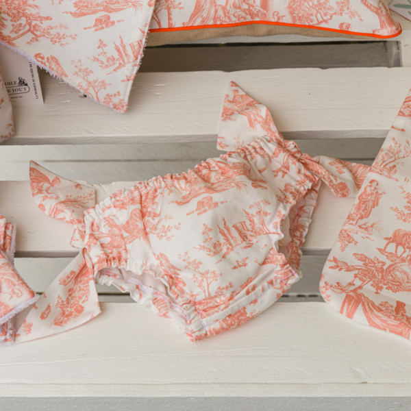 La culotte bouffante XCOMM_TROUSSE PENELOPE OFFRANDE PEACH PINK, ornée de volants rose pêche et blancs ainsi que de délicats nœuds, est présentée sur un support en bois blanc, aux côtés d'accessoires en tissu à motifs assortis. la marque Toile de Jouy