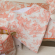 Gros plan sur le tissu XCOMM_TROUSSE PENELOPE OFFRANDE PEACH PINK, mettant en valeur sa forme triangulaire et ses motifs pastoraux de toile de corail représentant des personnages et des animaux sur fond blanc. D'autres articles en tissu aux motifs similaires sont partiellement visibles à proximité. la marque Toile de Jouy