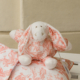Une « XCOMM_TROUSSE PENELOPE OFFRANDE PEACH PINK », composée d’un lapin en peluche aux longues oreilles et d’un tissu à motifs rose pêche, repose sur un coussin assorti posé sur une surface en bois blanc, devant un mur beige. la marque Toile de Jouy