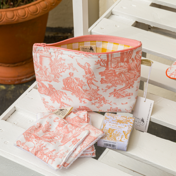 La pochette XCOMM_TROUSSE PENELOPE OFFRANDE PEACH PINK — une pochette rose et blanche à motif « toile » — repose sur un banc blanc, à côté de linges assortis et d’une petite boîte à savon décorative, avec une plante en pot visible à l’arrière-plan. la marque Toile de Jouy