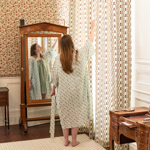 Une femme portant le XCOMM_KIMONO FLORAL SAUGE se tient pieds nus dans une pièce de style ancien au papier peint fleuri et au mobilier en bois, et tend la main pour toucher les rideaux assortis. Son reflet apparaît dans un miroir à cadre en bois à côté d'elle. la marque Toile de Jouy