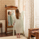 Une femme portant le XCOMM_KIMONO FLORAL SAUGE se tient pieds nus dans une pièce de style ancien au papier peint fleuri et au mobilier en bois, et tend la main pour toucher les rideaux assortis. Son reflet apparaît dans un miroir à cadre en bois à côté d'elle. la marque Toile de Jouy